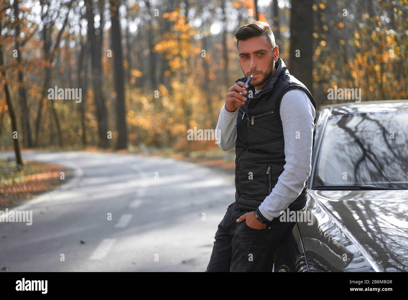 Un homme caucasien portant un gilet noir de style debout pres de la voiture noire sur la nature. Les jeunes hommes d affaires adultes voyagent en voiture sur une belle autu ensoleillee Photo
