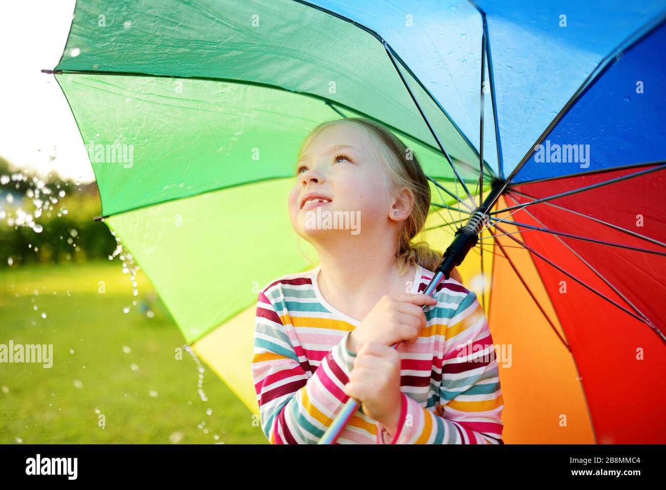 Jolie jeune fille tenant un parapluie arc-en-ciel coloré le jour de l'été  pluvieux. Enfant marchant sous la pluie chaude à l'extérieur. Activités  d'été en plein air pour les enfants Photo Stock -