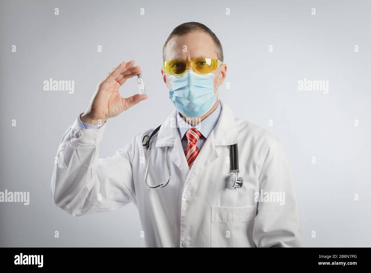 Le médecin montre le vaccin dans une capsule. Spécialiste médical dans une robe médicale, des lunettes et un masque et un stéthoscope sur ses épaules Banque D'Images