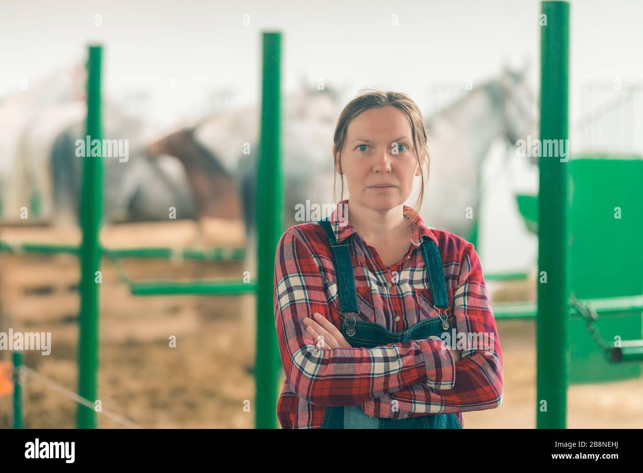 Portrait de femme au ranch horse stable looking at camera. Femme adulte portant des jeans et chemise à carreaux salopette comme ouvrier agricole. Banque D'Images