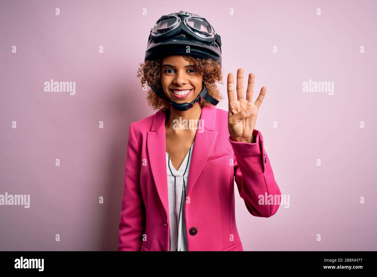 Femme automobiliste afro-américaine avec des cheveux bouclés portant un casque moto sur fond rose montrant et pointant vers le haut avec les doigts numéro quatre tandis que sm Banque D'Images