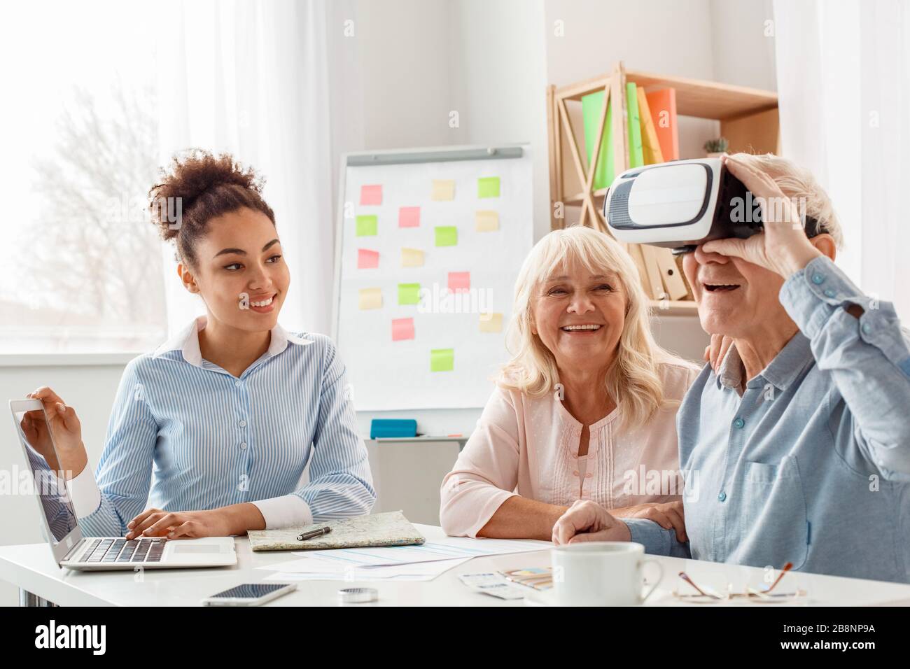 Agent de voyage au bureau utilisant un ordinateur portable regardant les clients seniors homme portant un casque de réalité virtuelle souriant excité Banque D'Images