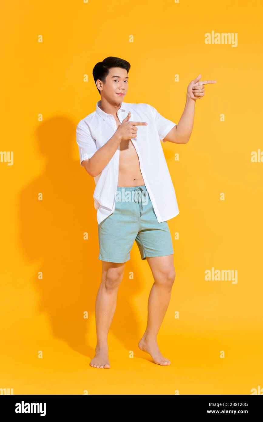 Corps entier de jeune homme asiatique dans un ensemble d'été décontracté pointant deux mains de côté sur fond jaune coloré Banque D'Images