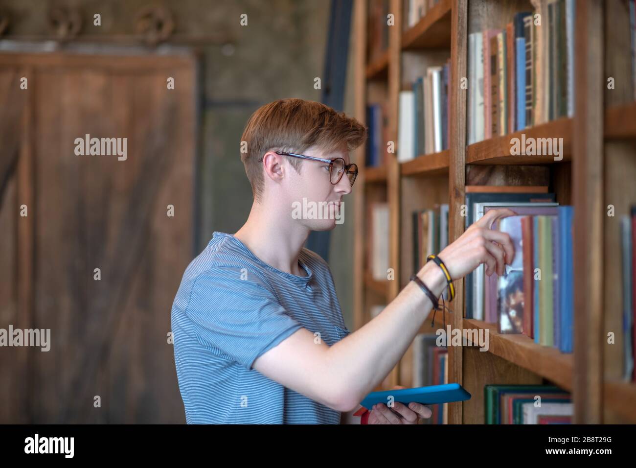 Jeune homme dans des lunettes prenant un livre d'une étagère de livre Banque D'Images