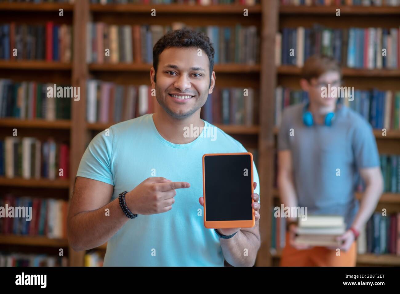 Jeune latino à poil foncé dans un t-shirt bleu montrant sa tablette Banque D'Images