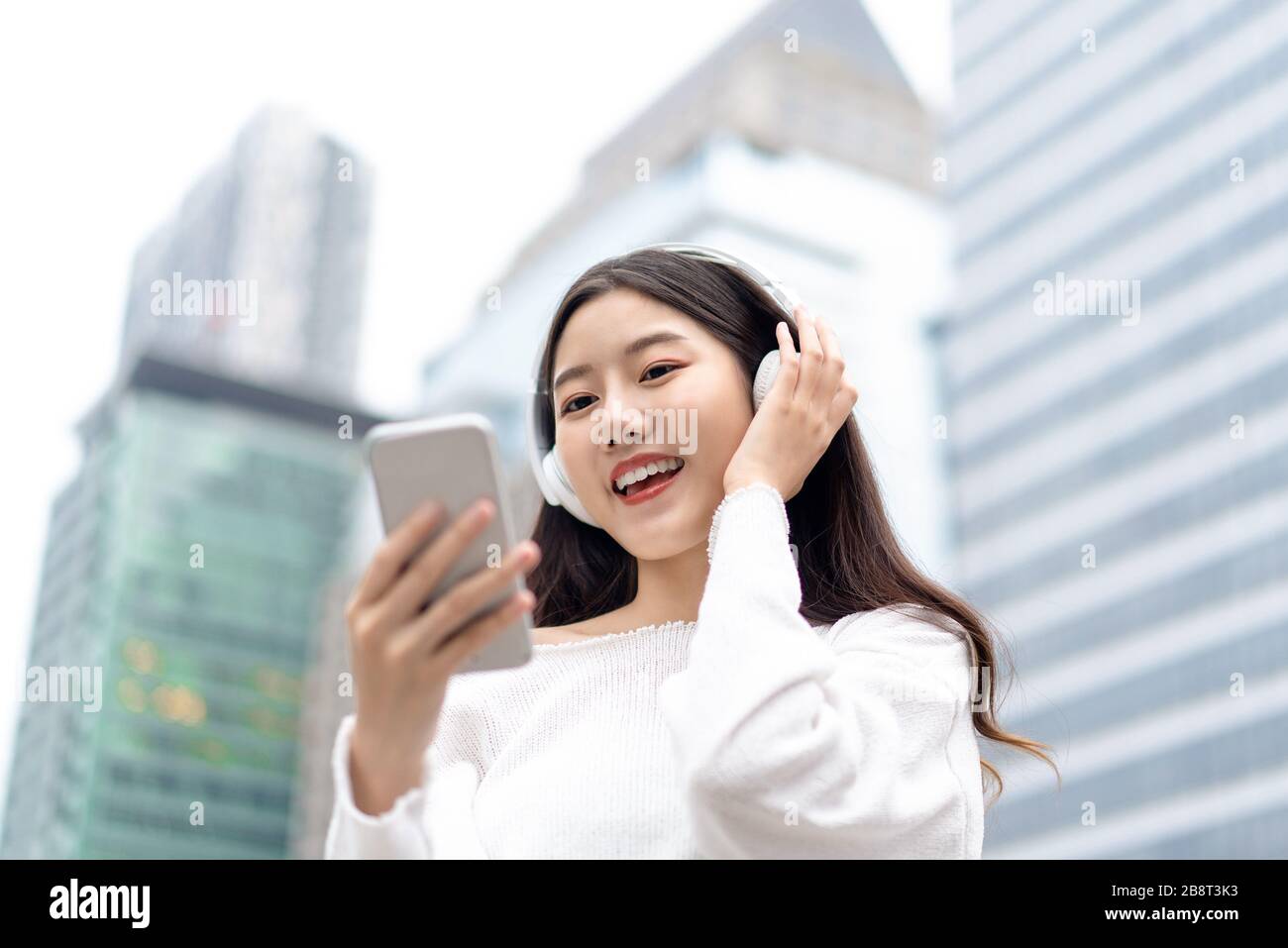 Heureuse femme asiatique souriante portant un casque et regardant le téléphone mobile tout en écoutant de la musique en streaming sur fond de bâtiment de ville Banque D'Images