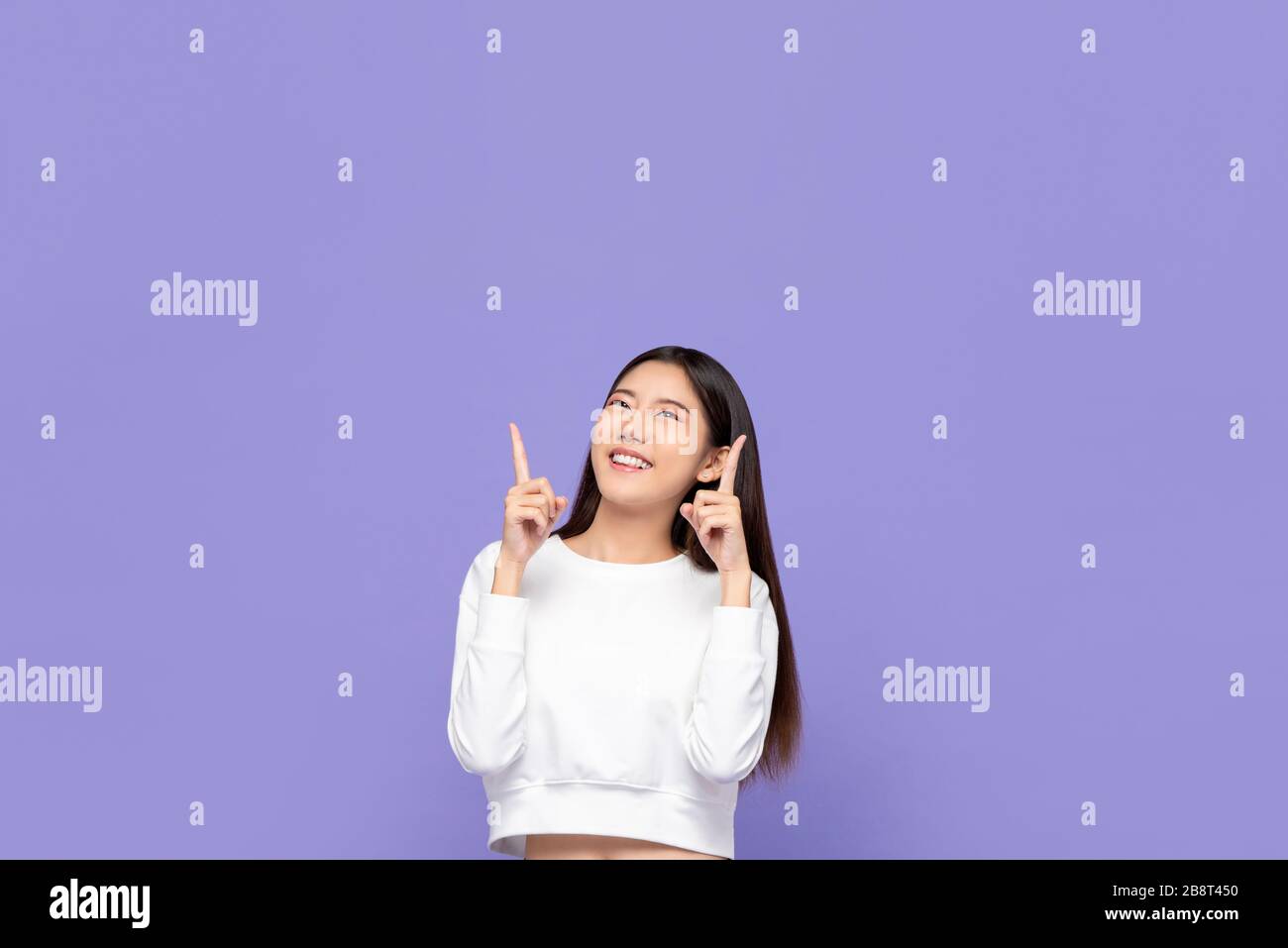 Portait d'une jeune femme asiatique souriante mignonne pointant vers le haut et regardant un espace vide au-dessus dans un fond de studio isolé pourpre Banque D'Images