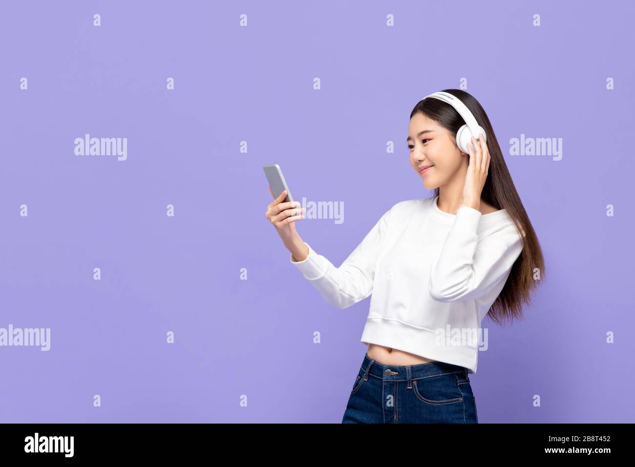 Portrait d'une jeune femme asiatique souriante écoutant de la musique sur un téléphone mobile à l'aide d'un casque sans fil dans un fond de studio violet isolé Banque D'Images