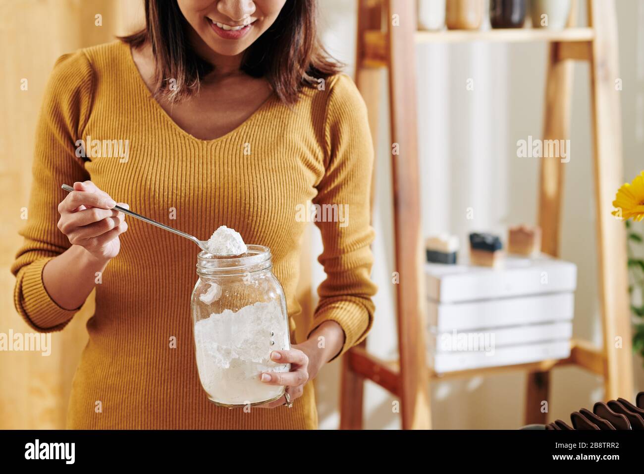 Jeune femme souriante prenant une cuillère de lessive de soda à partir d'un pot en verre lors de la fabrication de savon parfumé à la maison Banque D'Images