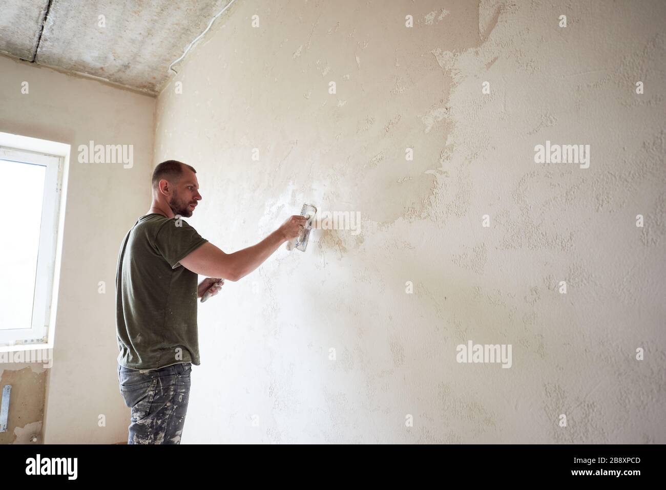 Le travailleur de l'homme fait la réparation dans l'appartement en mettant la putty sur les murs. Jeune homme avec une barbe dans un t-shirt et un short enduit de peinture travaille jour. Maison de construction. Banque D'Images