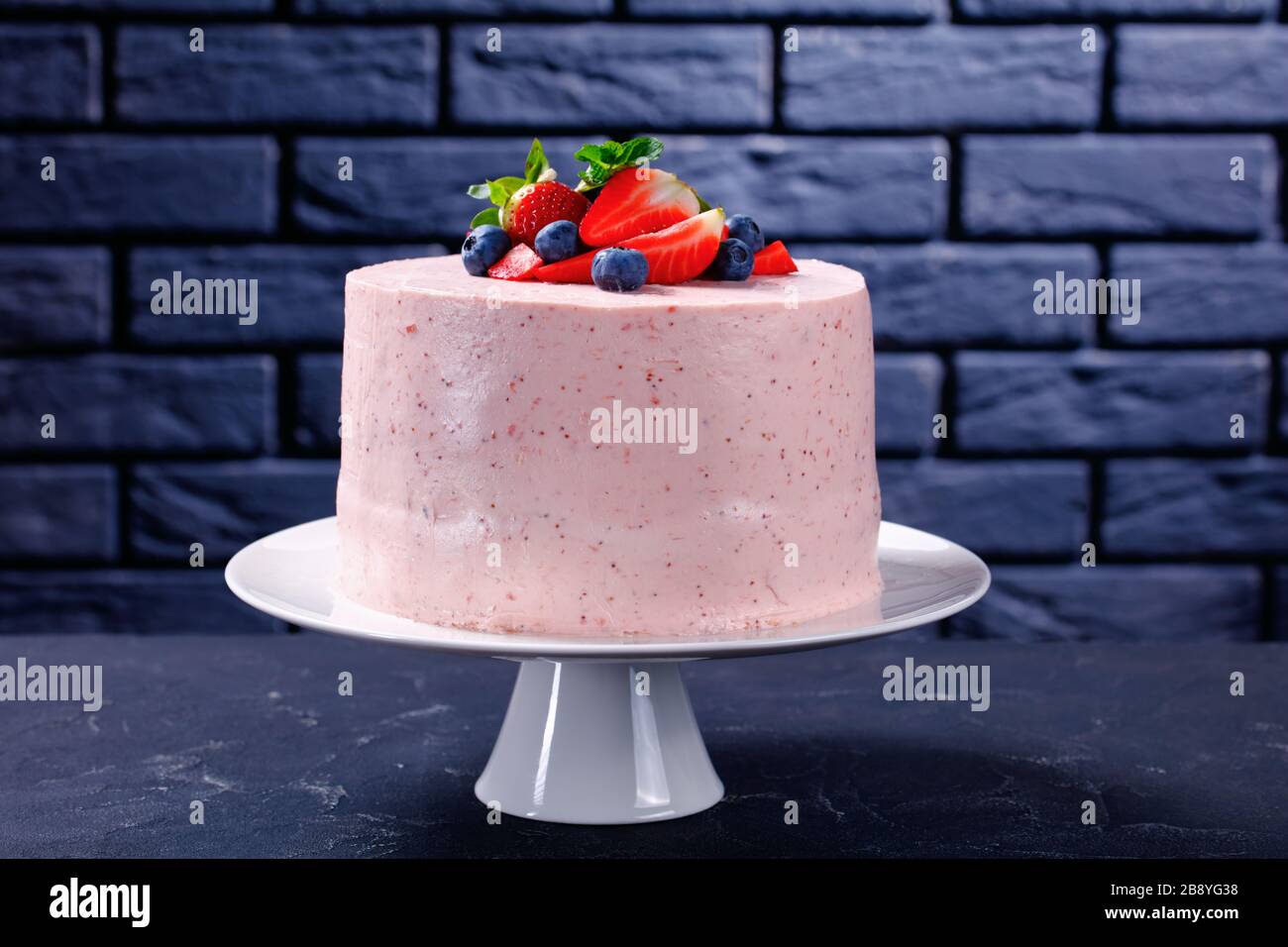 Delicieux Et Sucre Un Gateau De Fraise Rose Entier Decore De Fruits Frais Et De Feuilles De Menthe Sur Un Stand De Gateau Blanc Avec Un Mur De Brique A L Arriere Plan Photo