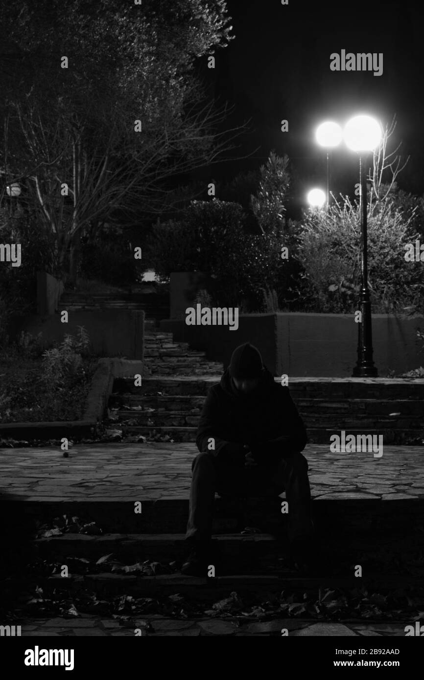 Homme assis dans un parc éclairé par des feux de rue la nuit. Noir et blanc. Banque D'Images