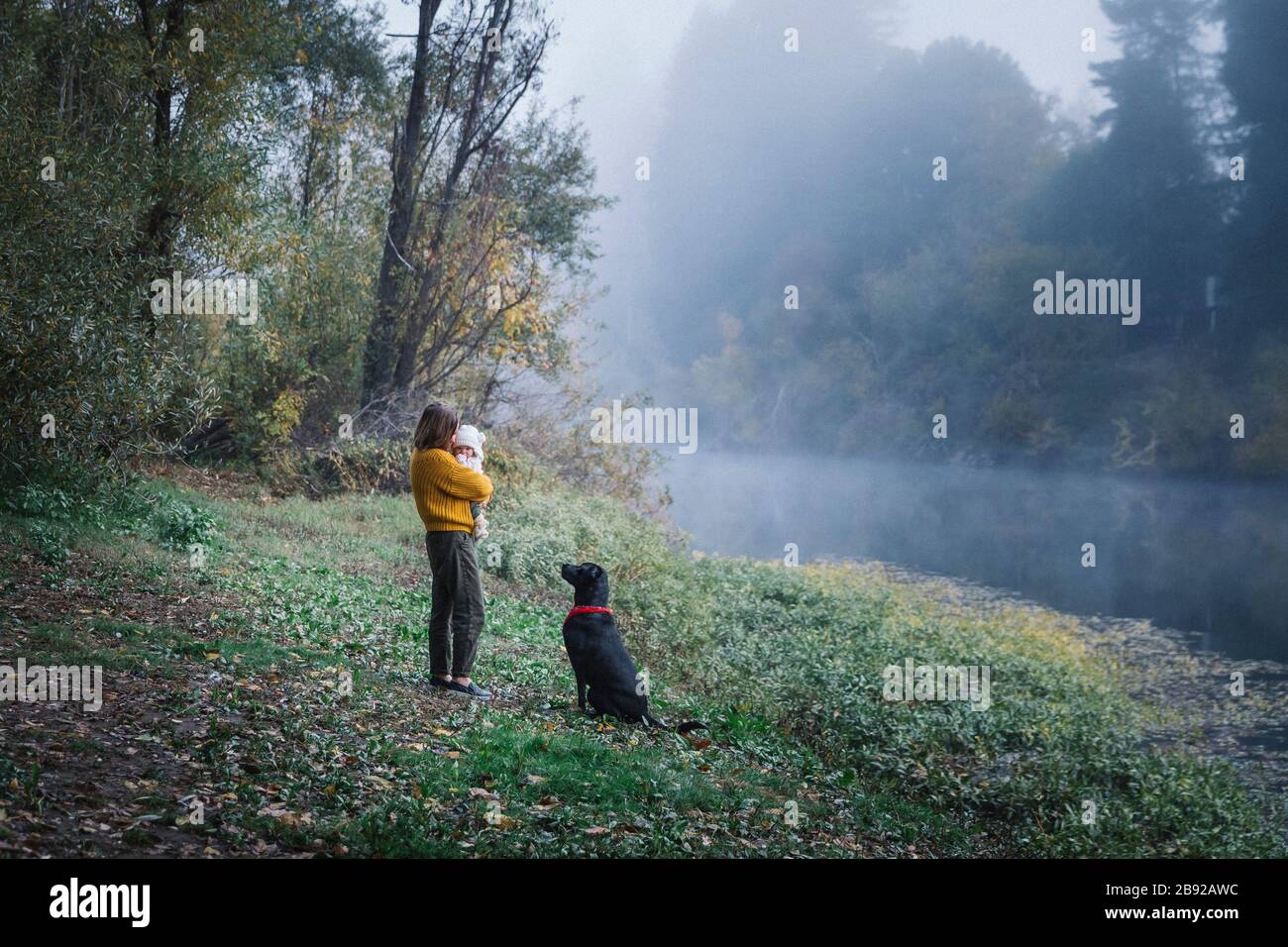 Une femme tient un bébé et regarde un chien près d'une rivière Banque D'Images