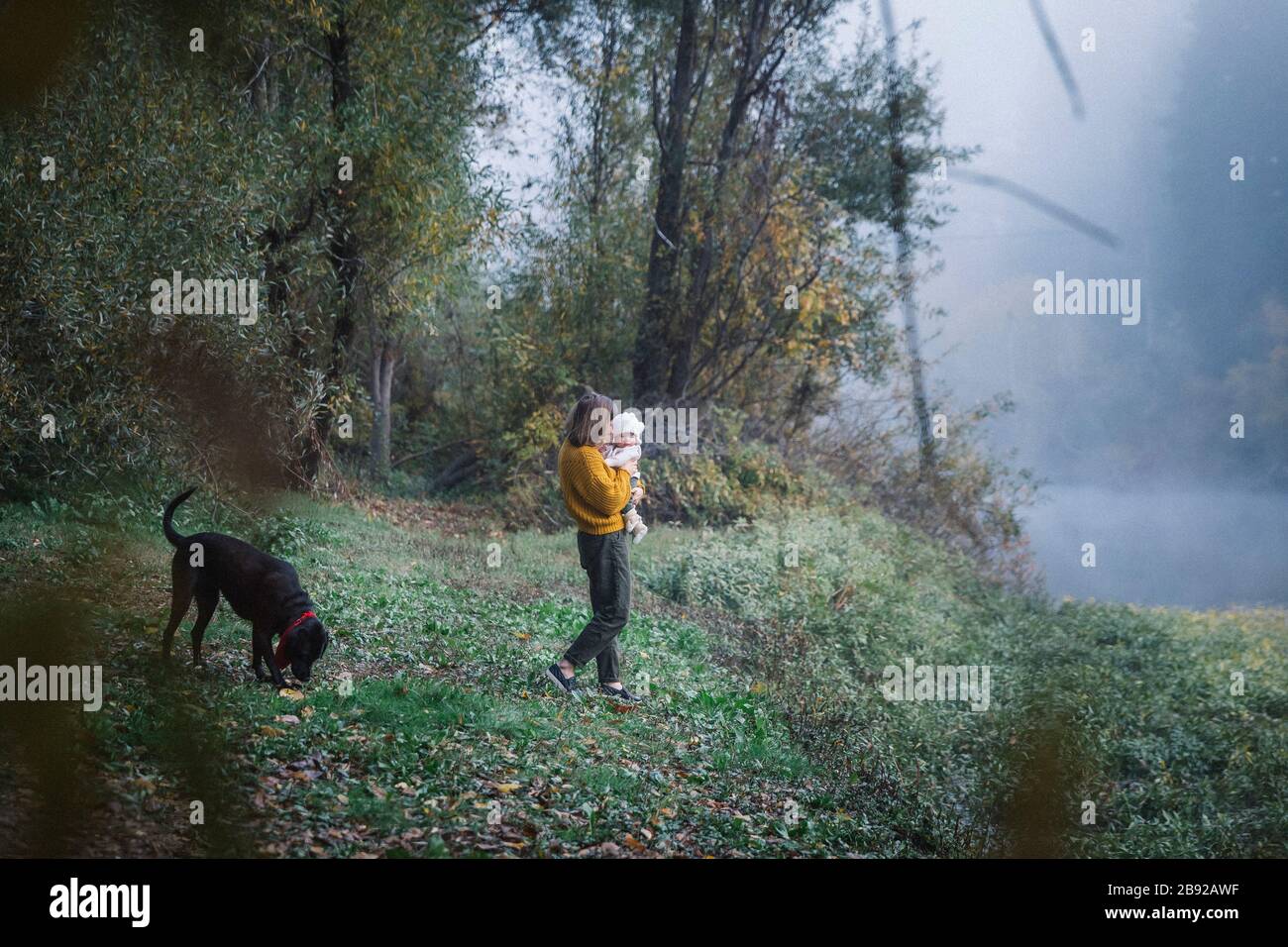 Une femme tient un bébé et regarde un chien près d'une rivière Banque D'Images