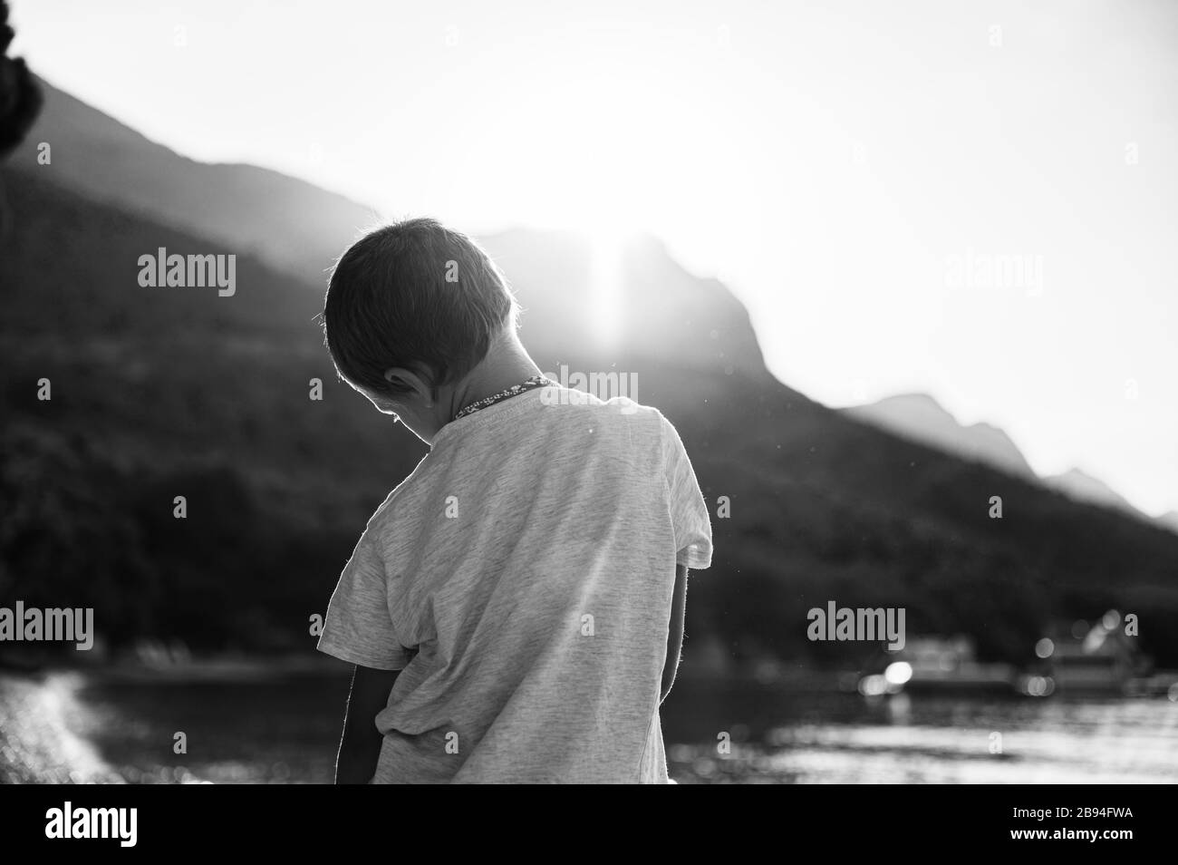 Vue de derrière un petit garçon se tenant dehors par la mer avec le soleil levant venant de derrière les collines. Image monochrome. Banque D'Images