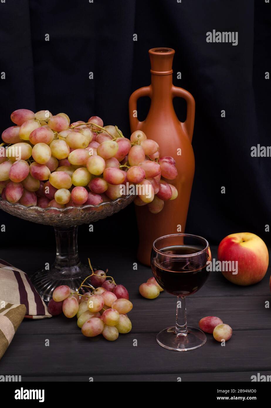Vin rouge dans un verre avec des raisins dans un vase sur une table en bois sur un fond de tissu noir avec une vieille bouteille. Pour la publicité du vin, la vie encore. Banque D'Images
