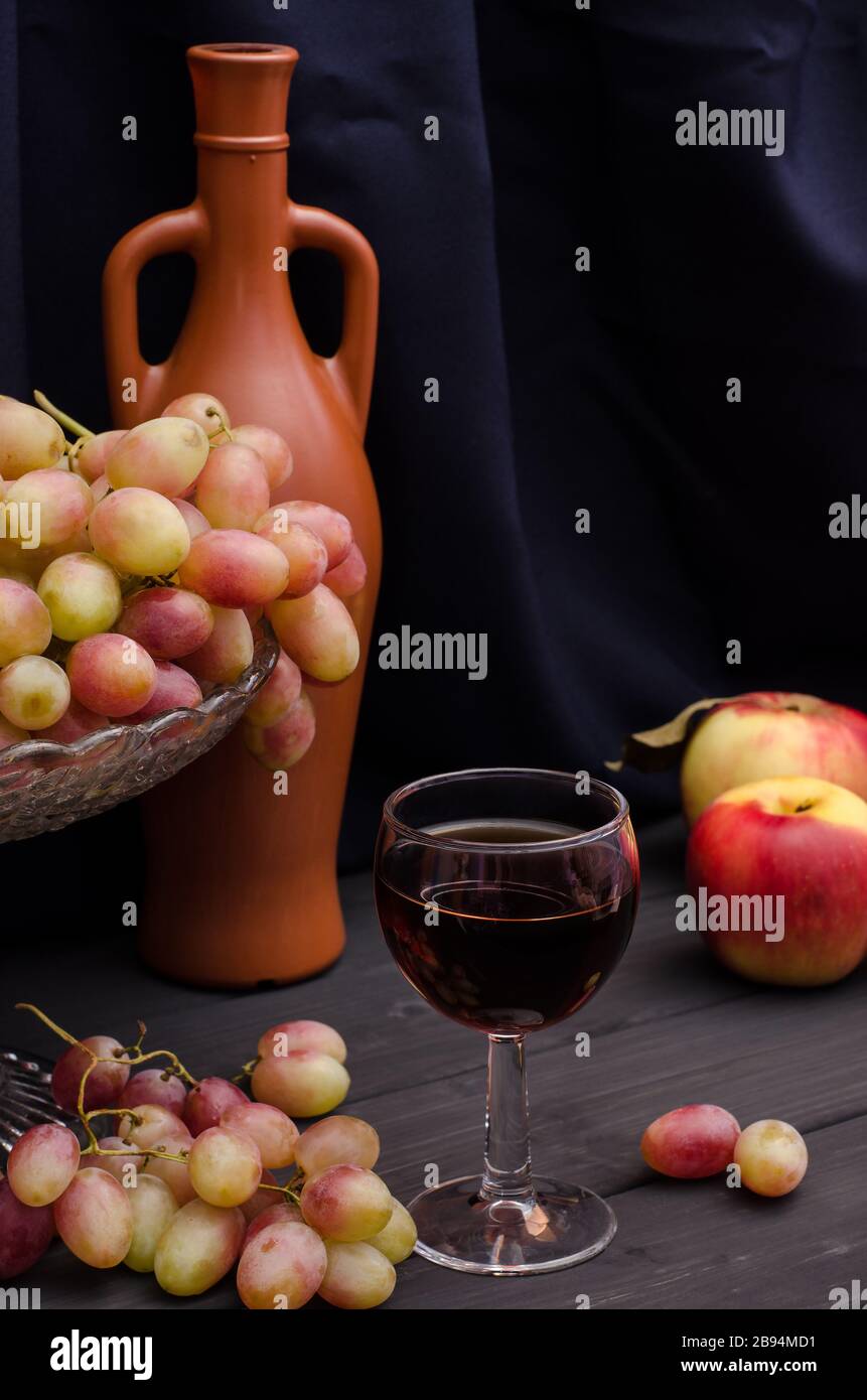 Vin rouge dans un verre avec des raisins dans un vase sur une table en bois sur un fond de tissu noir avec une vieille bouteille. Pour la publicité du vin, la vie encore. Banque D'Images