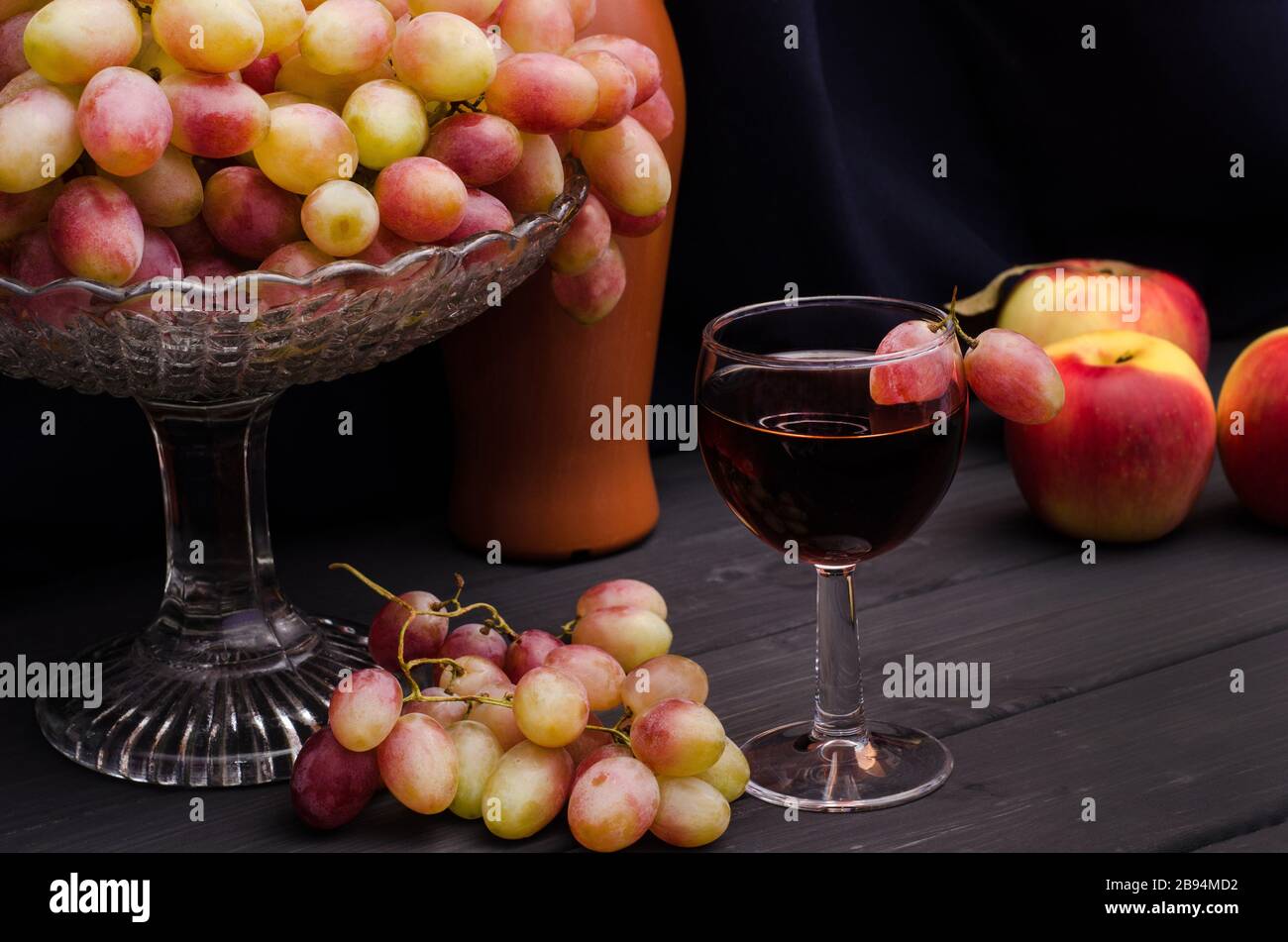 Vin rouge dans un verre avec des raisins dans un vase sur une table en bois sur un fond de tissu noir avec une vieille bouteille. Pour la publicité du vin, la vie encore. Banque D'Images