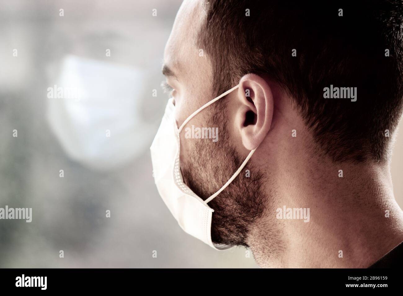 Photo d'un jeune homme blanc avec masque de visage médical regardant hors de la fenêtre pendant la quarantaine du coronavirus. Reflet de son visage dans la fenêtre. Coronavirus, éclosion de COVID-19. Concept médecin/infirmière. Banque D'Images