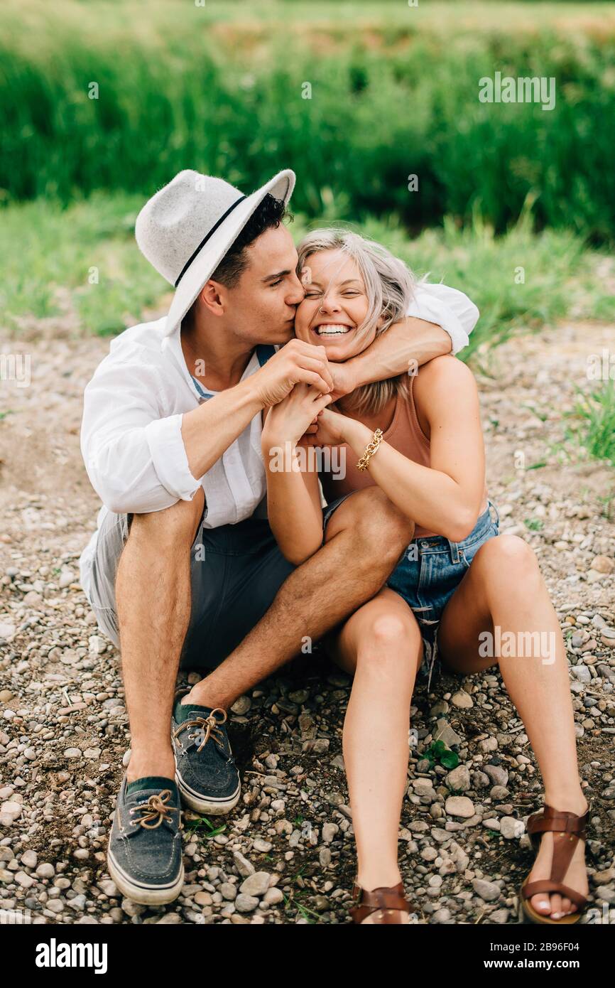 Jeune couple assis sur une rive, souriant et embrassant. Banque D'Images