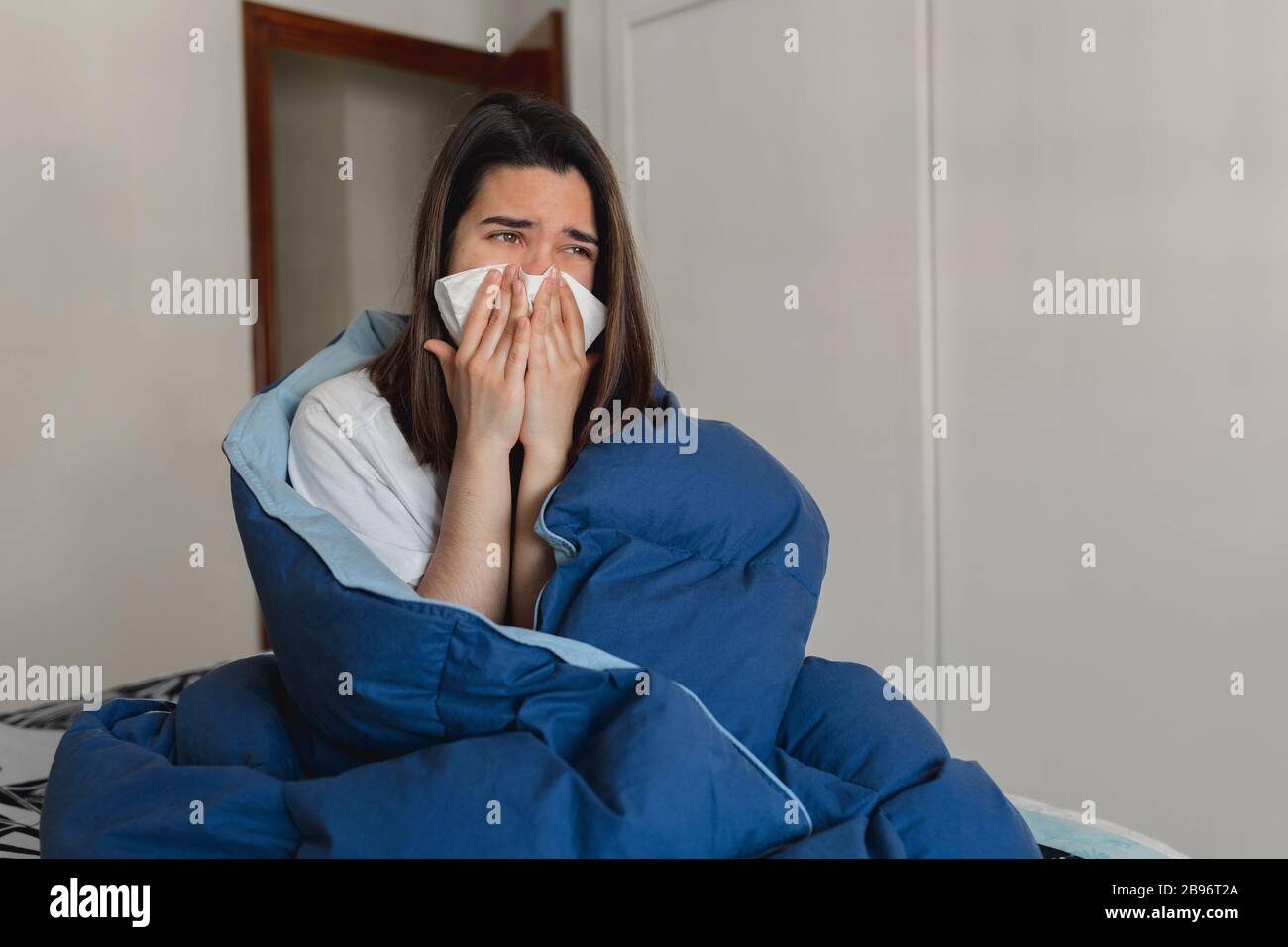 Jeune femme malade éternuant dans un tissu tout en couvrant avec une quilt bleu dans sa maison Banque D'Images