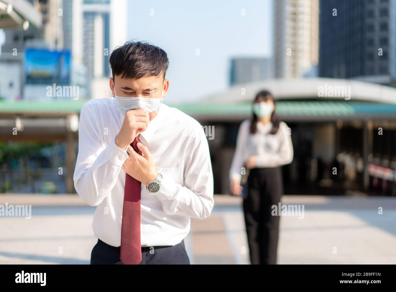 Un homme d'affaires asiatique malade tousse avec masque avec femme d'affaires en arrière-plan pour maintenir la distance de protection contre les virus COVID-19 et les personnes social distancing FO Banque D'Images