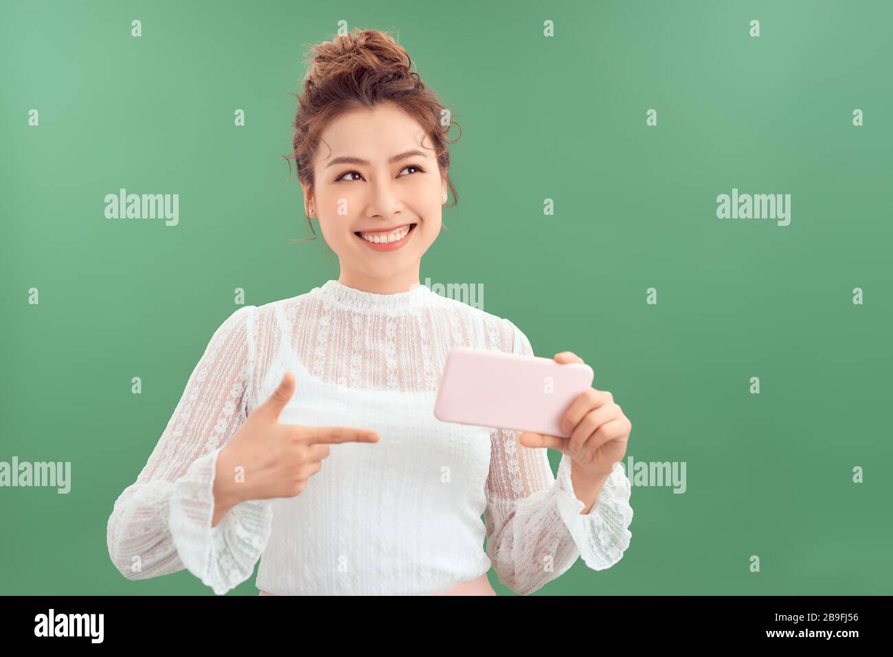 Belle jeune femme asiatique montrant le téléphone mobile tout en se tenant et en regardant isolé sur fond vert. Banque D'Images