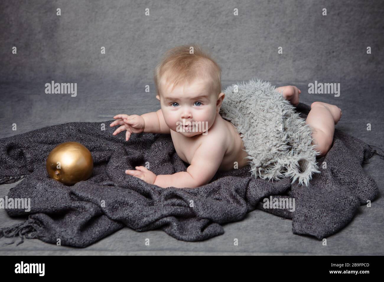 Une petite fille allongée sur son front sur une couverture grise douce, prise sur un fond gris de studio, elle regarde la caméra. Banque D'Images