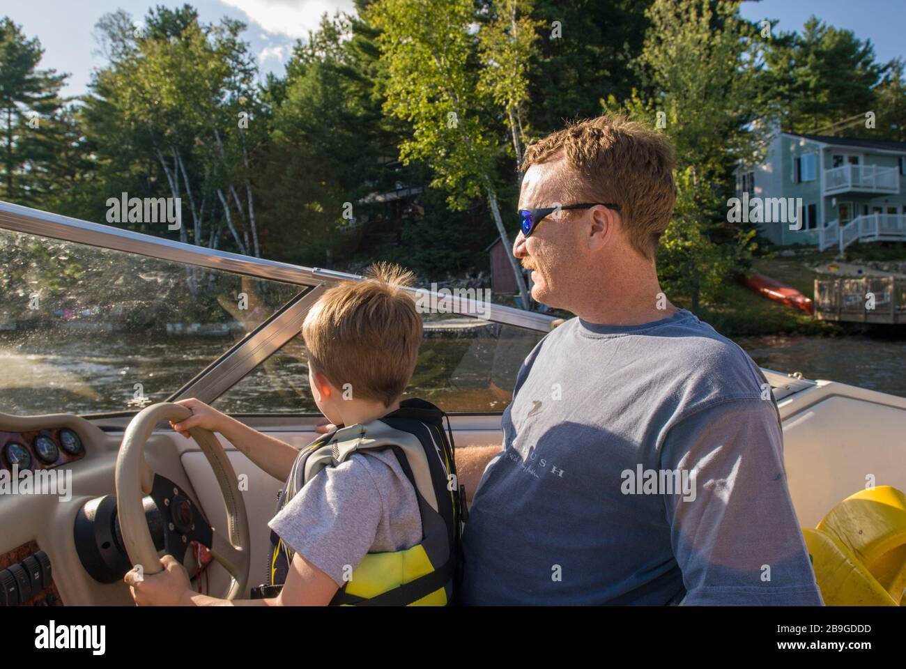 Garçon de 7 ans en gilet de sauvetage assis sur les genoux de son père à la roue d'un bateau à moteur sur Balch Pond à Wakefield, New Hampshire, à la fin de l'été Banque D'Images