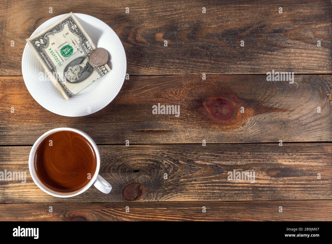 Tasse blanche avec café et soucoupe espresso revigorants avec deux billets et pièces de monnaie américains. Sur fond en bois sombre. Paiement ou conseil. Vue de dessus. CO Banque D'Images