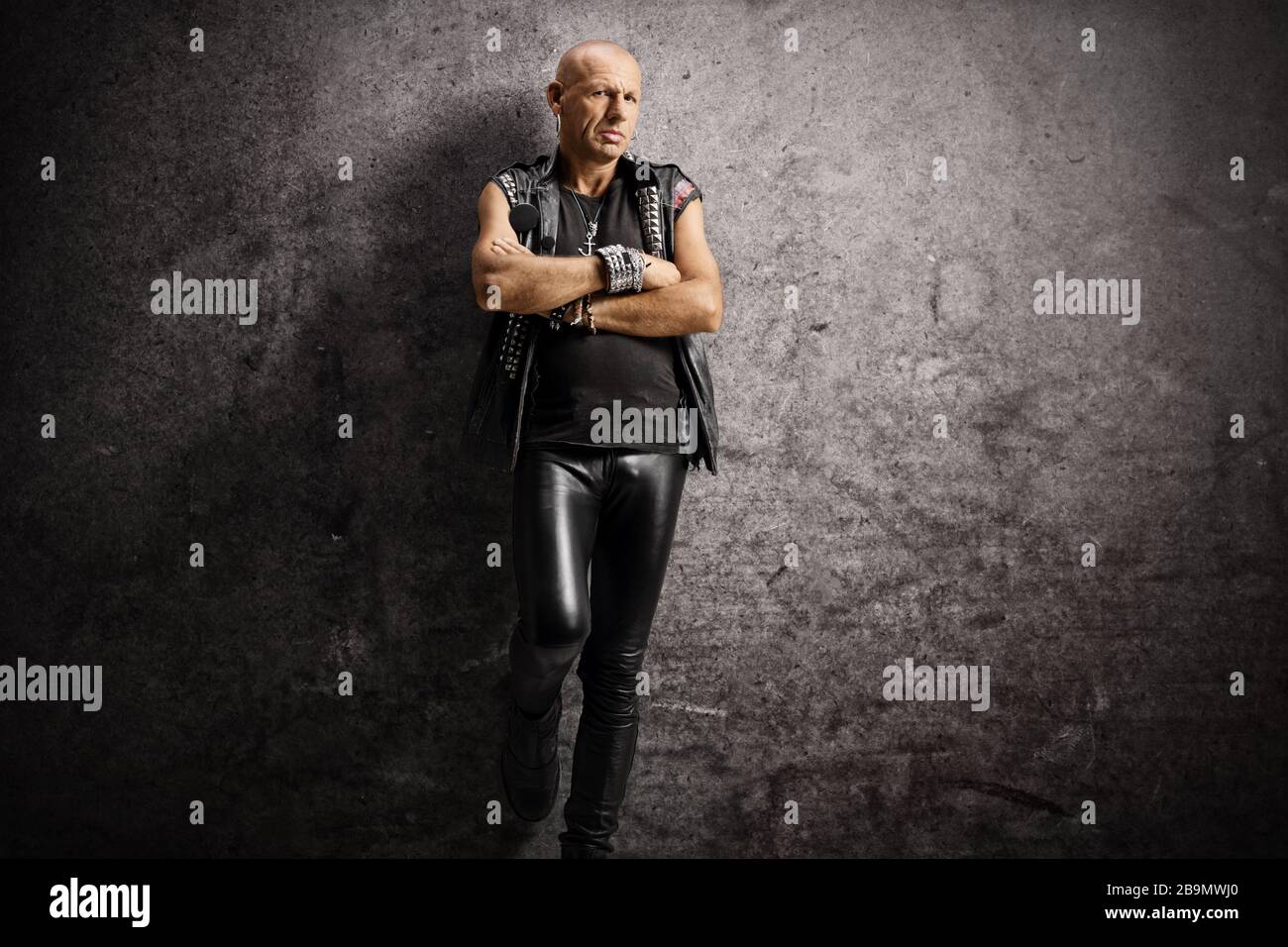 Un punk chauve sérieux penchant sur un mur rouillé et posant avec des bras croisés Banque D'Images