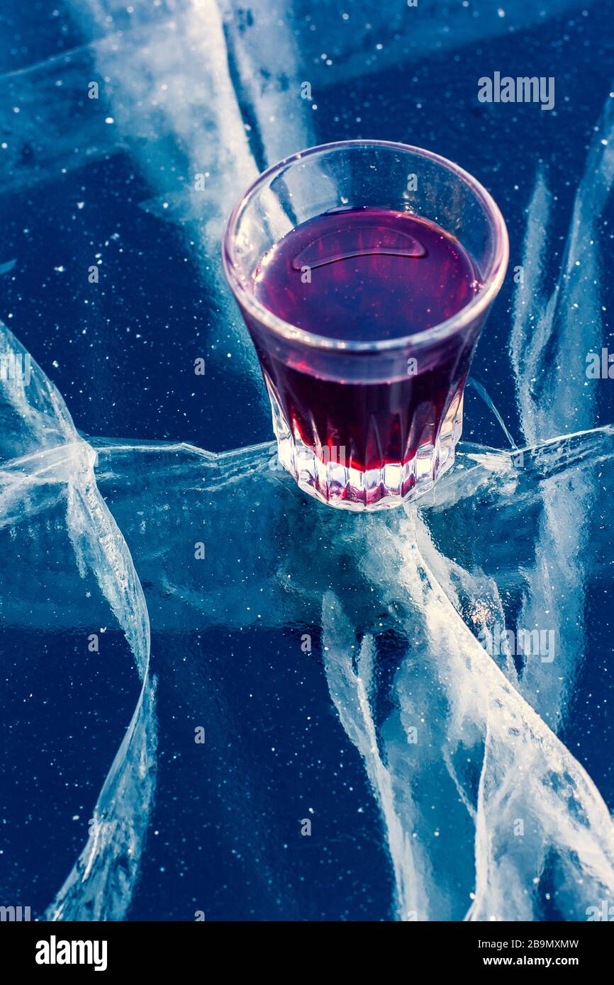 L'alcool dans un verre et nettoyer la glace avec de belles fissures profondes. Un verre avec teinture rouge se dresse sur la glace transparente du lac. Vertical. Banque D'Images