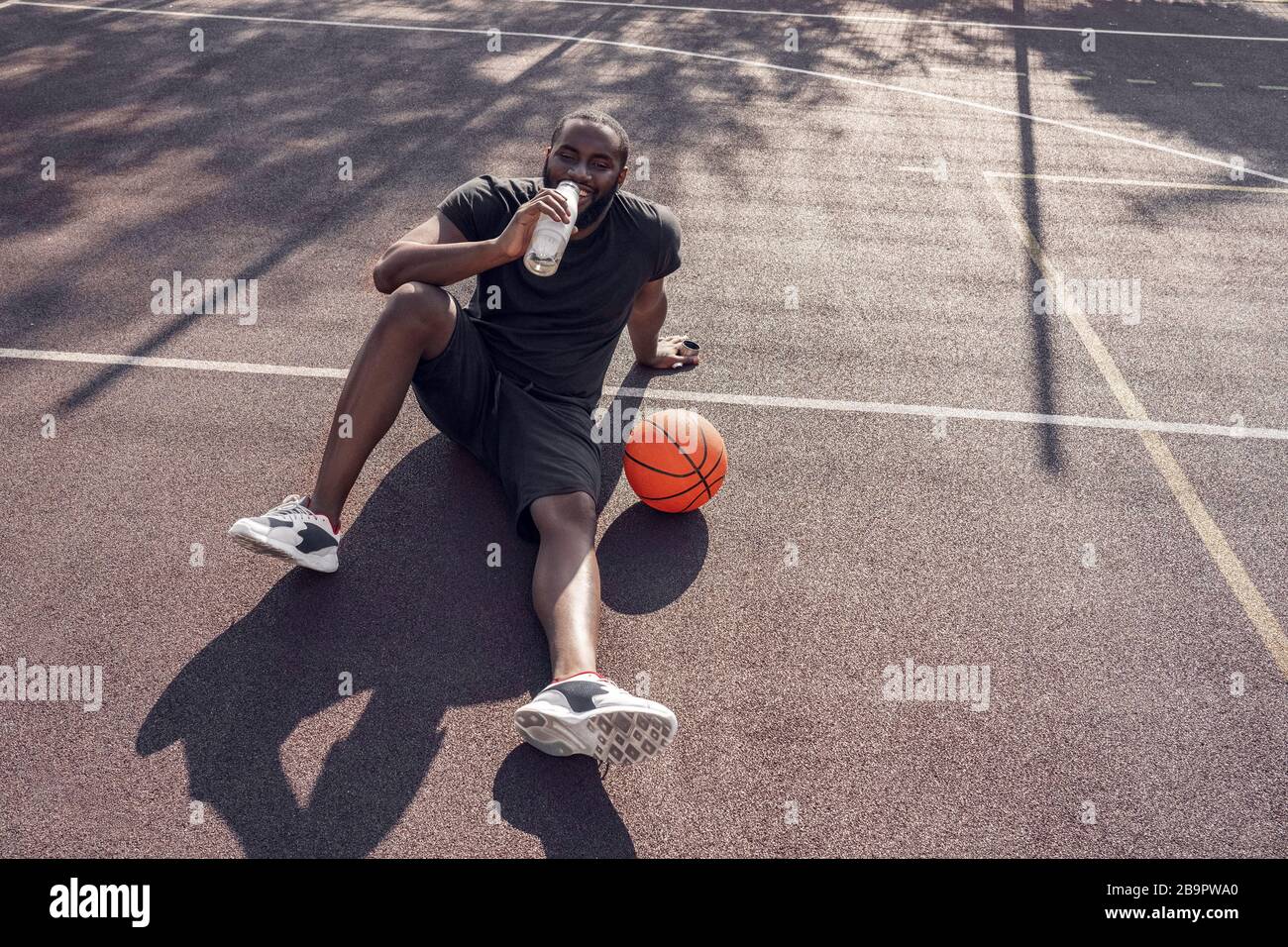 Activités en plein air. Homme africain assis sur le terrain de basket-ball avec de l'eau potable de balle souriant détendu Banque D'Images