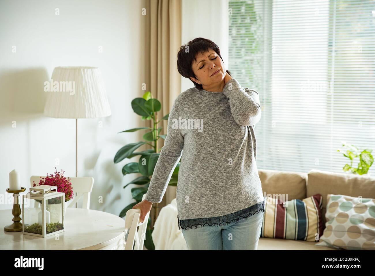 Femme mûre souffrant de mal au dos à la maison. Masser le cou avec la main, se sentir épuisé, debout dans le salon. Banque D'Images