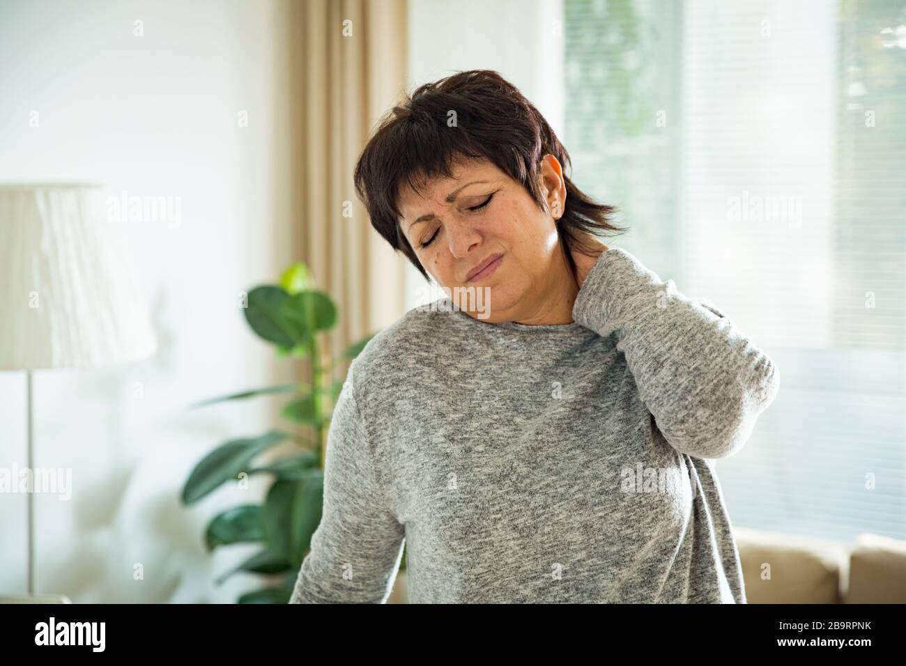 Femme mûre souffrant de mal au dos à la maison. Masser le cou avec la main, se sentir épuisé, debout dans le salon. Banque D'Images