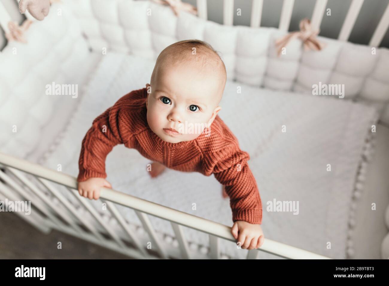 Belle petite fille dans un lit de bébé dans une salle de lumière confortable dans un pull de tons à la mode. Apprend à se tenir au soutien. Petite fille gaie debout dans le lit de bébé à la maison Banque D'Images