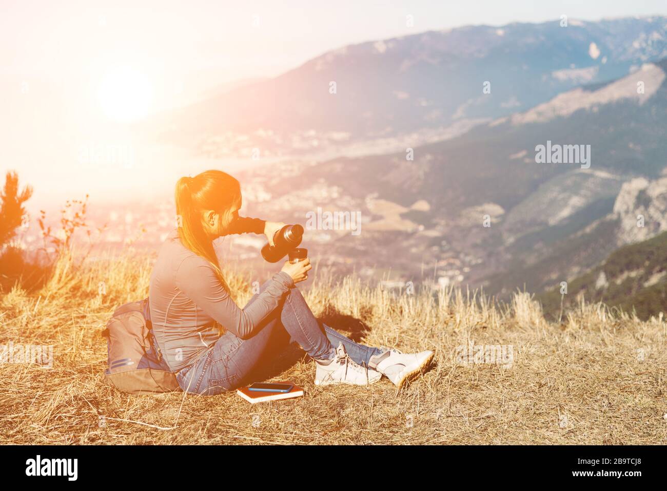 La jeune fille se déplace seule dans les montagnes au printemps ou en automne, se trouve au bord de la montagne et regarde la distance et jouit de la nature, des rochers et des forêts vertes, vue sur le paysage. Un sac à dos derrière et vêtements de sport, un thermos avec une boisson chaude ou du thé, liberté et légèreté. Vue arrière de la femme voyageur en casquette assis seul sur le sommet de la montagne et en regardant le beau paysage d'été et la vue mer bleue. Buvez du thé ou du café Banque D'Images