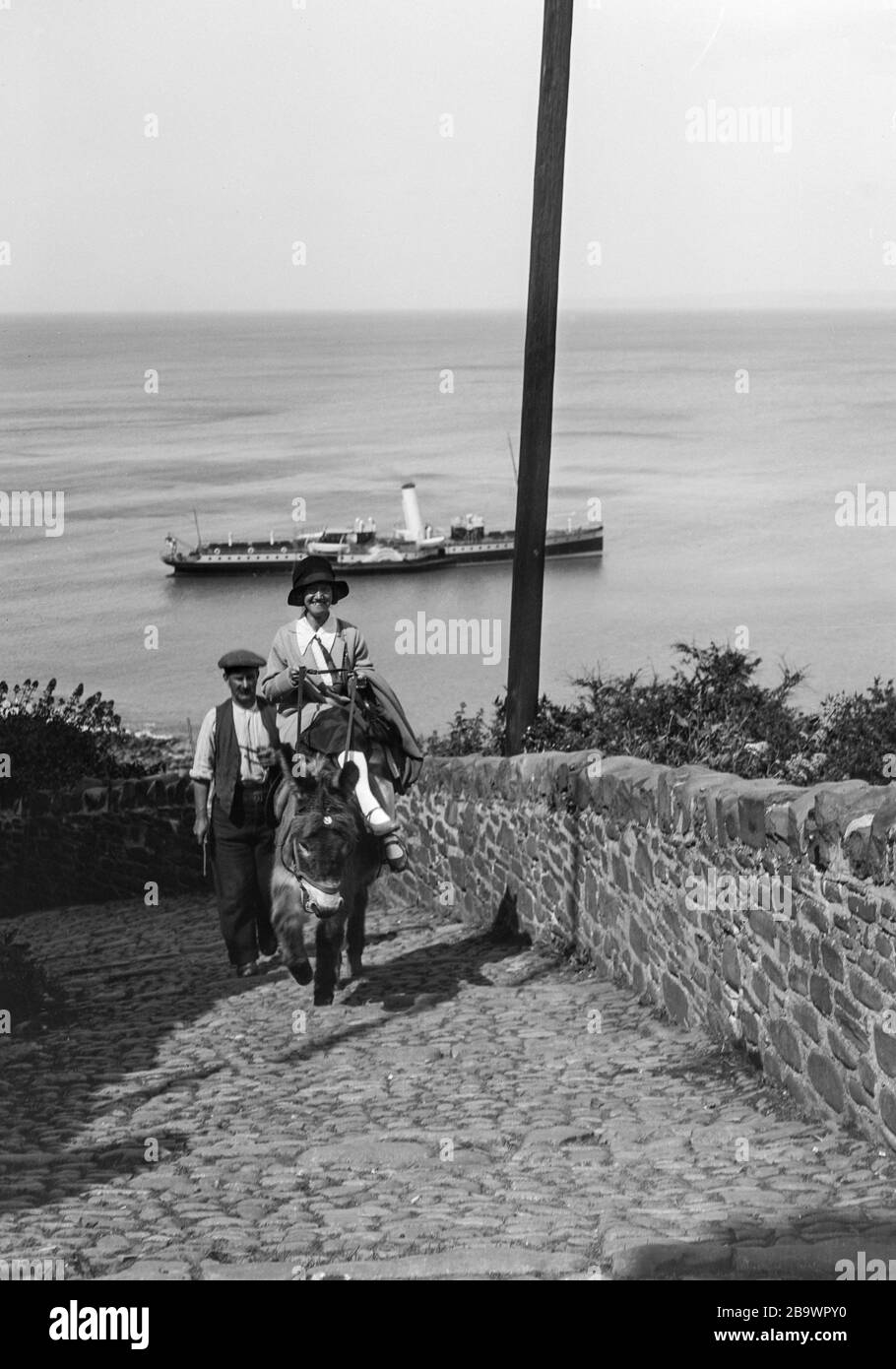 Image d'archive vers 1925 d'une femme qui monte un âne de la plage au village de Clovelly, Devon, Royaume-Uni, montrant un défroisseur en arrière-plan. Numérisé à partir du négatif d'origine. Banque D'Images