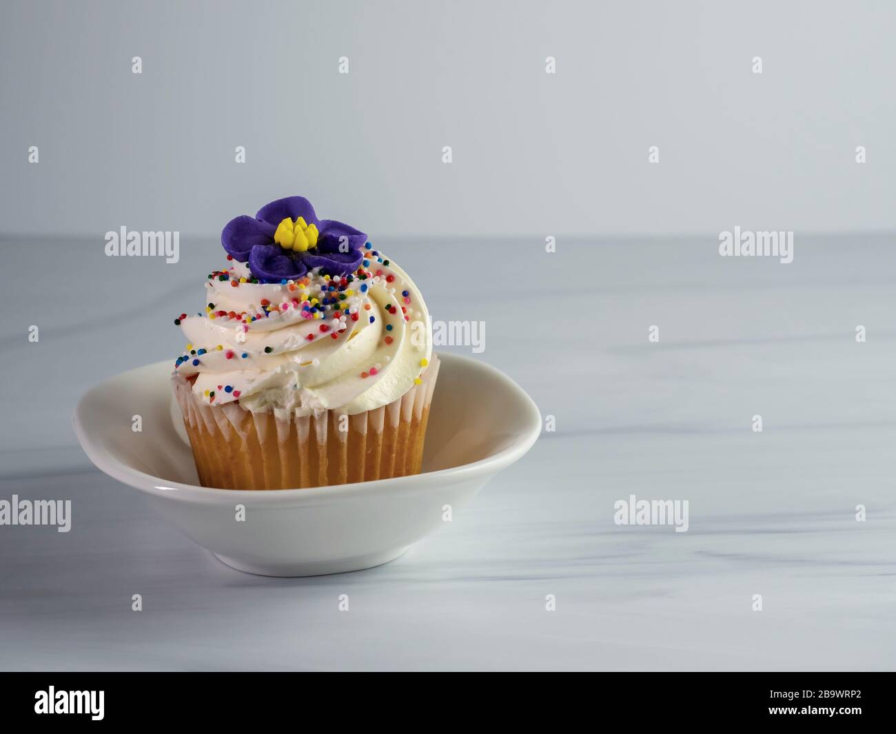 Gâteau à la vanille avec girled blanc girrant, une fleur de fondant pourpre et des arroseurs assis dans un plat blanc en céramique sur un comptoir en grès, délicieux A Banque D'Images