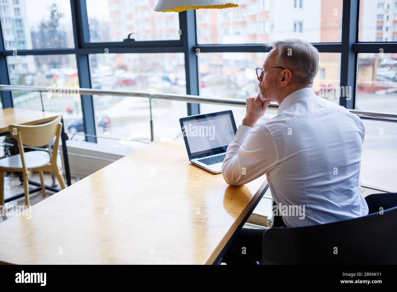 Le travailleur indépendant travaille dans un café sur un nouveau projet d'affaires. Se trouve sur une grande fenêtre à la table. Regarde un écran d'ordinateur portable avec une tasse de café Banque D'Images