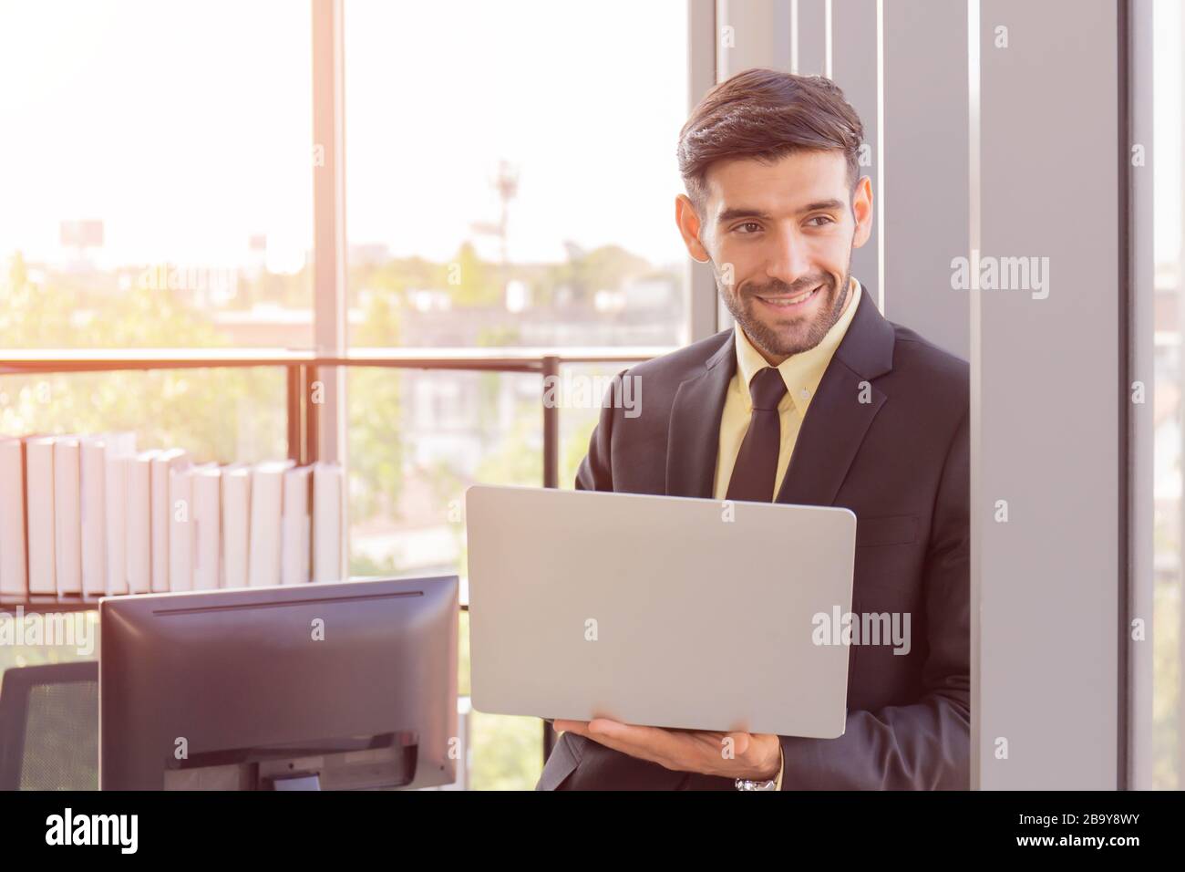 Un homme d'affaires habillé d'un costume bien habillé tient une tablette à côté d'un mur de verre dans son bureau avec un sourire brillant et une belle lumière orange. Banque D'Images