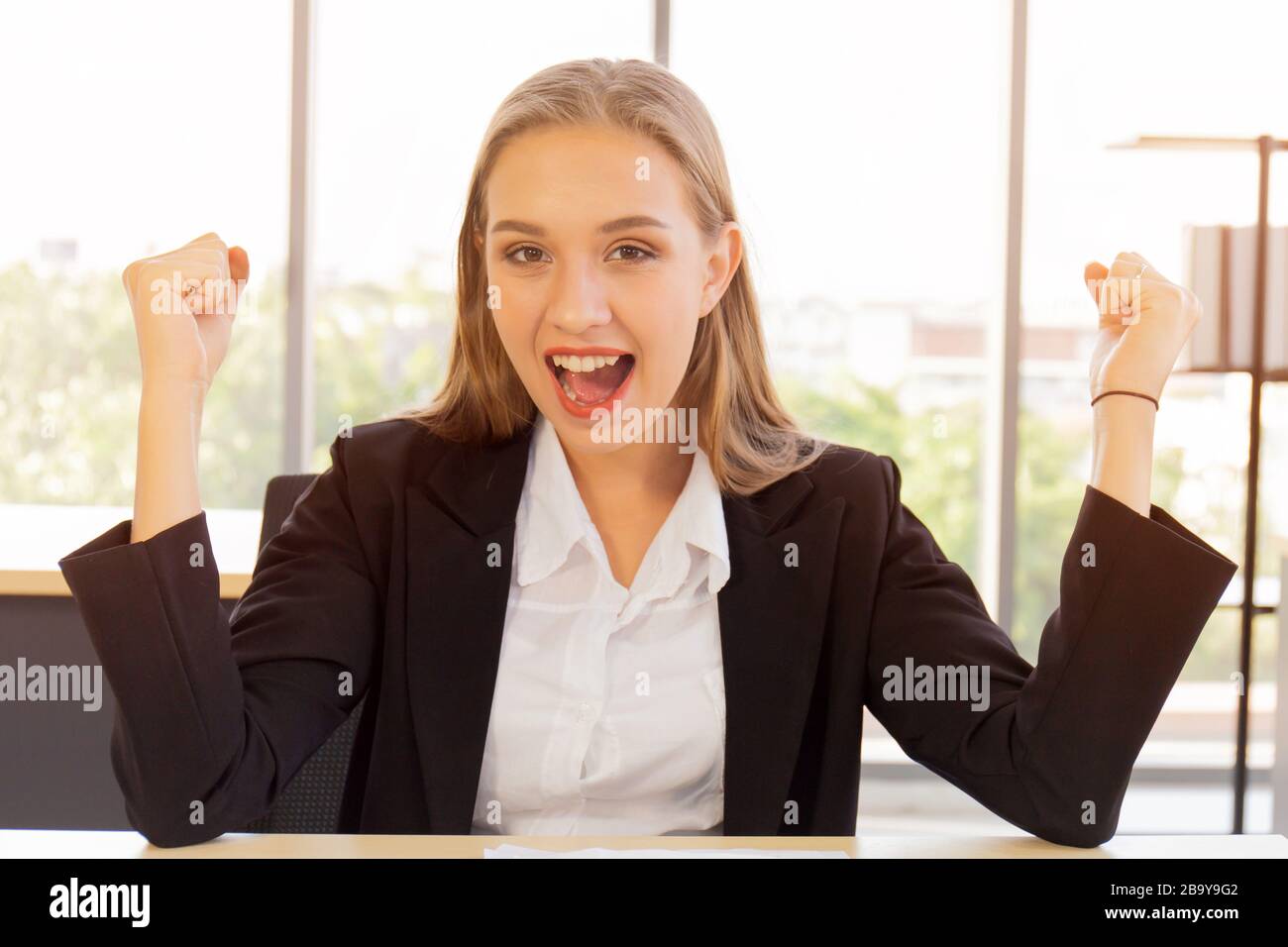 Une belle femme d'affaires en costume, bien habillée, assise au bureau, très heureuse, a levé la main avec un sourire vif sur la table Banque D'Images