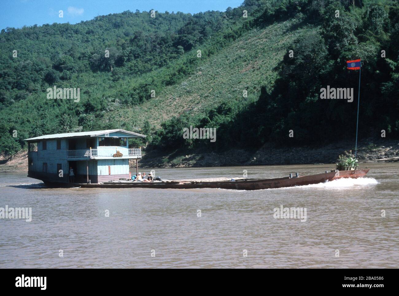 Les touristes voyagent comme passagers de pont sur un bateau lent le long du Mékong au Laos, entre Huay Xai et Luang Prabang. L'équipage vit et travaille dans les quartiers en bois de la poupe sur l'artisanat. À la prow se trouve un drapeau lao et des pots de fleurs roses. Un champ a été éliminé dans la jungle sur la banque derrière. Banque D'Images