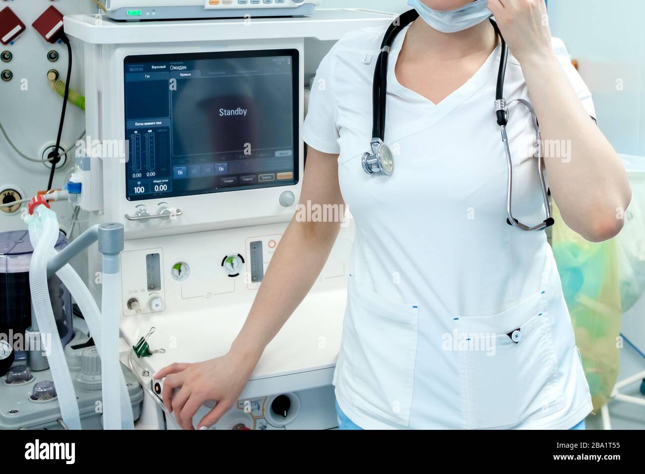 Un anesthésiste en vêtements médicaux blancs, un masque et un stéthoscope sur son cou se trouvent dans la salle d'opération. Derrière le médecin se trouvent des moniteurs de hig Banque D'Images