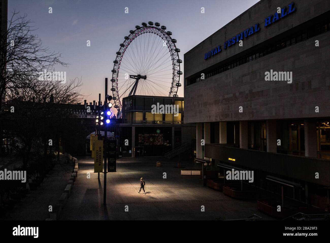 Londres, Royaume-Uni. 25 mars 2020. Une figure solitaire illuminée par des lumières de rue traverse une salle de festival royale vide au crépuscule alors que le « verrouillage de Coronavirus » du West End de Londres continue de faire face à la propagation rapide de la maladie dans toute la capitale. Les Economiistes prévoient également un krach économique dû aux restrictions et les fermetures de théâtres, de restaurants et de commerces pourraient coûter plus de vie que Cronavirus. Crédit : Jeff Gilbert/Alay Live News Banque D'Images