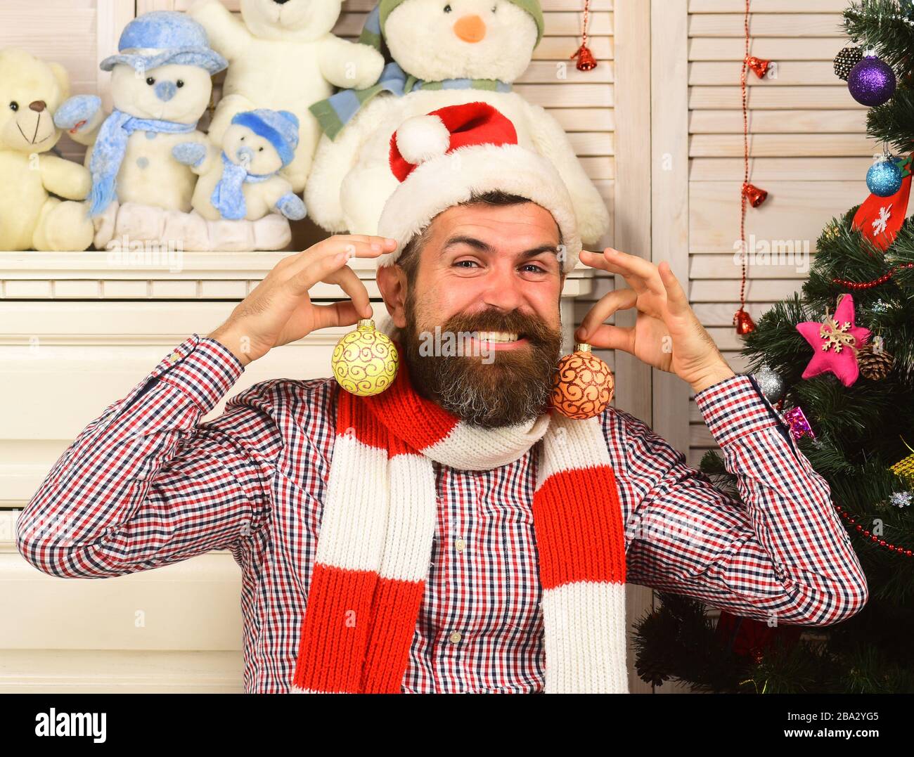 Concept de festivals et de décor. Guy fait des décorations. L'homme avec  barbe tient des boules de Noël dorées comme boucles d'oreilles. Père Noël  avec visage heureux avec mur en bois, bureau