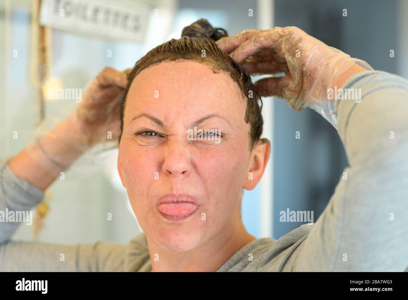 Femme tirant un visage et se collant de sa langue comme elle applique la teinture de cheveux à ses cheveux brun frisé à la maison dans un gros portrait Banque D'Images