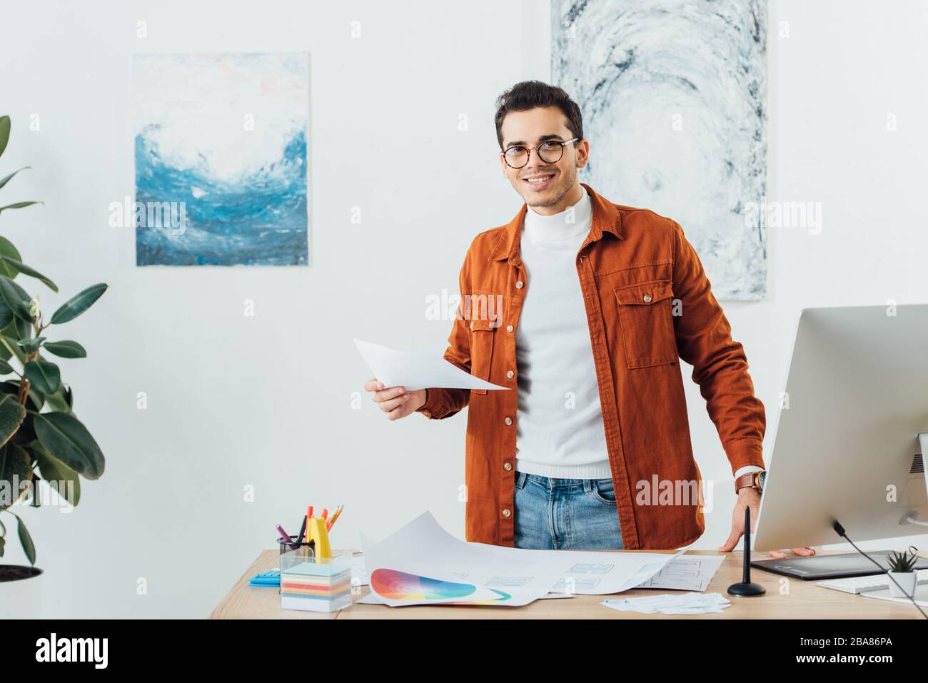 Concepteur souriant regardant l'appareil photo tout en travaillant avec des cercles de couleur et des croquis de site Web à la table Banque D'Images