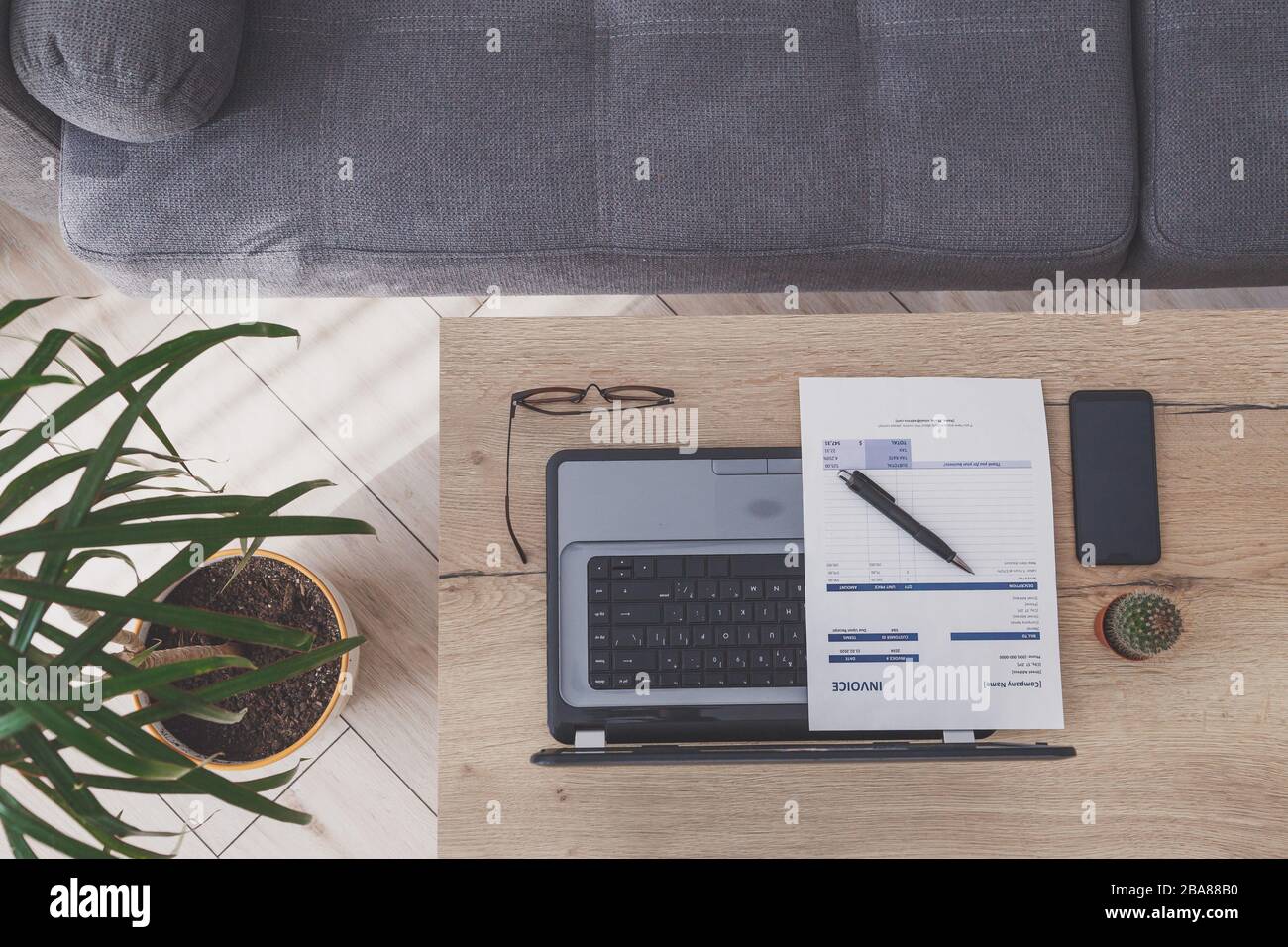 Vue de dessus du lieu de travail du bureau à domicile. Ordinateur portable, facture et lunettes sur table en bois, canapé Banque D'Images
