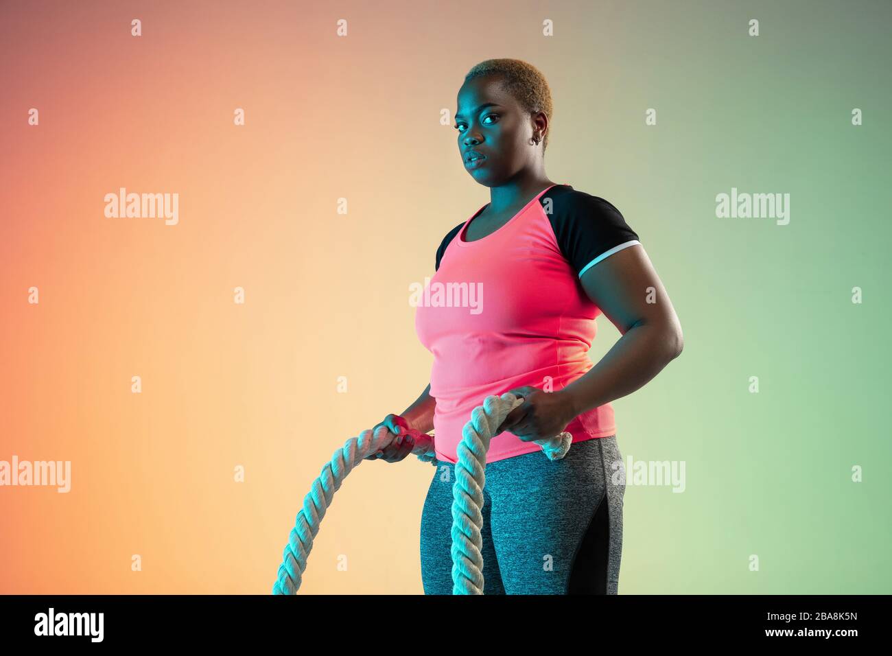 La formation des jeunes afro-américaines et des femmes de taille plus sur fond dégradé dans la lumière néon. Faire des exercices d'entraînement avec les cordes. Concept de sport, mode de vie sain, corps positif, égalité. Banque D'Images